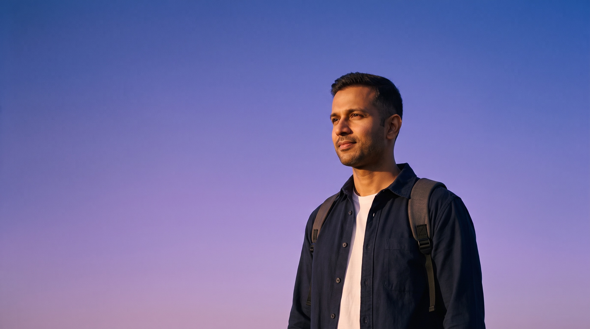 A man in a navy shirt with a backpack looks toward the evening light against a violet sky.